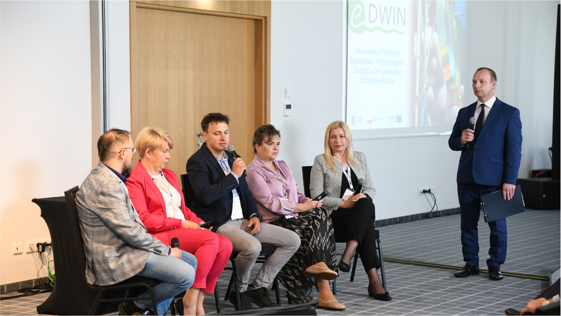 Grupa pięciu osób siedzi na krzesłach wewnątrz sali konferencyjnej. To uczestnicy panelu dyskusyjnego. Obok nich z mikrofonem w ręku stoi moderator w garniturze.