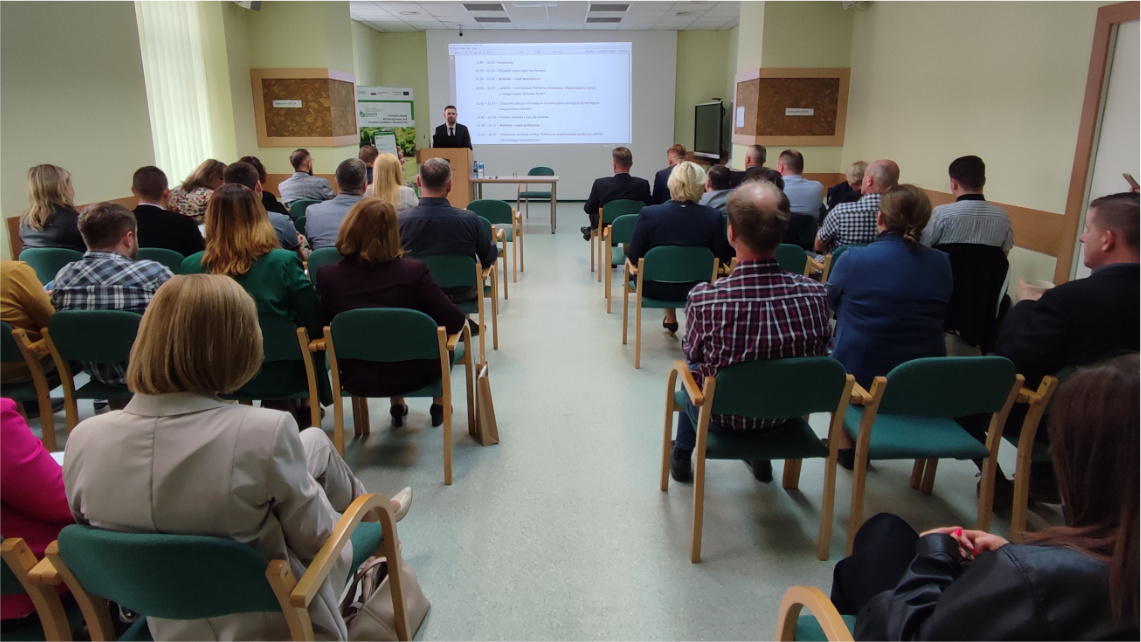 Wnętrze pomieszczenia. Na zdjęciu widać od tyłu grupę kilkudziesięciu osób siedzących na krzesłach podczas konferencji oraz patrzących na prezentację na rzutniku, która jest przed nimi.