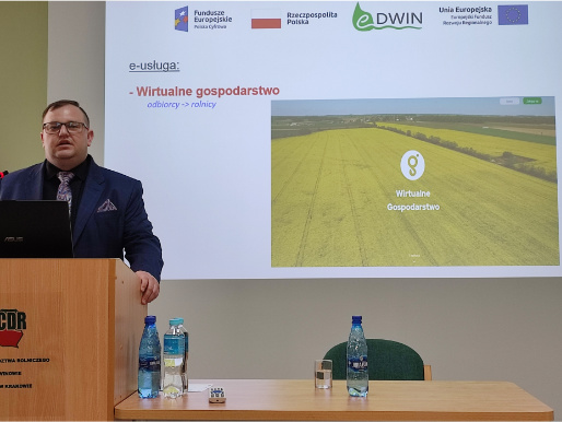 Michał Głombicki z Wielkopolskiego Ośrodka Doradztwa Rolniczego w Poznaniu opowiadał o praktycznym wykorzystaniu usługi Wirtualnego gospodarstwa Wnętrze pomieszczenia. Michał Głombicki, młody mężczyzna, stoi w marynarce przed rzutnikiem, na którym widoczna jest prezentacja o wirtualnym gospodarstwie. Przed nim jest mównica, na której stoi laptop.